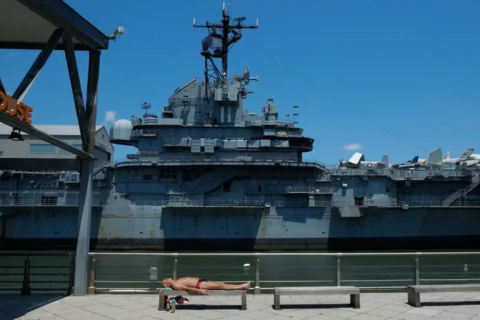 A man sunbathes in his small bathing trunks on a bench near the Intrepid